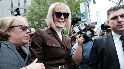 E Jean Carroll smiles, surrounded by reporters and wearing sunglasses, as she leaves a courthouse.