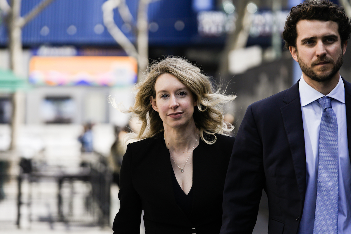 A young blonde woman (Elizabeth Holmes) looks into the camera directly in front of her while walking down a city street with a man.