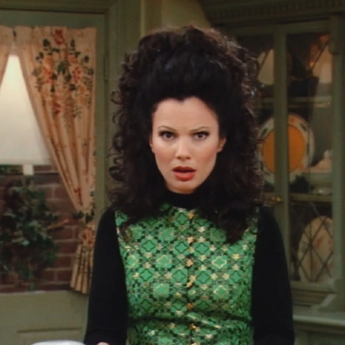 Image of Fran Drescher as Fran Fine in the CBS sitcom, 'The Nanny.' She is standing in the house looking right into the camera with a shocked expression on her face. Fran is a white, Jewish woman with long, curly, black hair worn half up in a ponytail and half down. She's wearing a green patterned top over a long-sleeved black shirt.