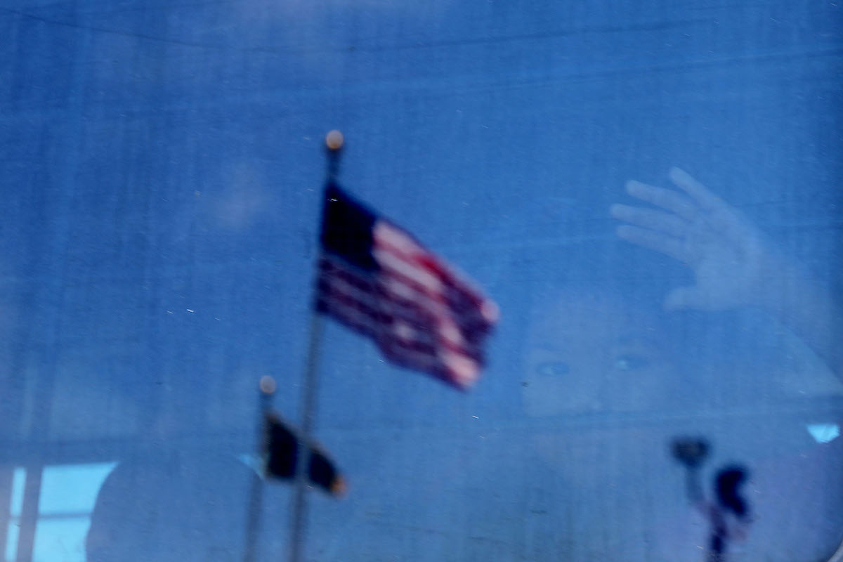 A child is vaguely seen through a window, with the focus on a reflection of an American flag flying outside.