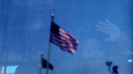 A child is vaguely seen through a window, with the focus on a reflection of an American flag flying outside.