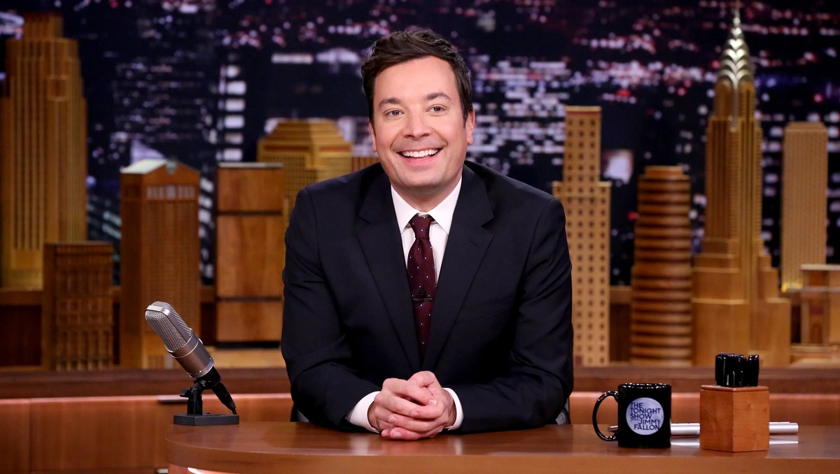 Image of Jimmy Fallon on the set of NBC's 'The Tonight Show.' He is a white man with short, dark hair wearing a black suit with a white buttondown shirt and a maroon tie with white dots on it. He's seated at a wooden desk with a background depicting the New York City skyline. 