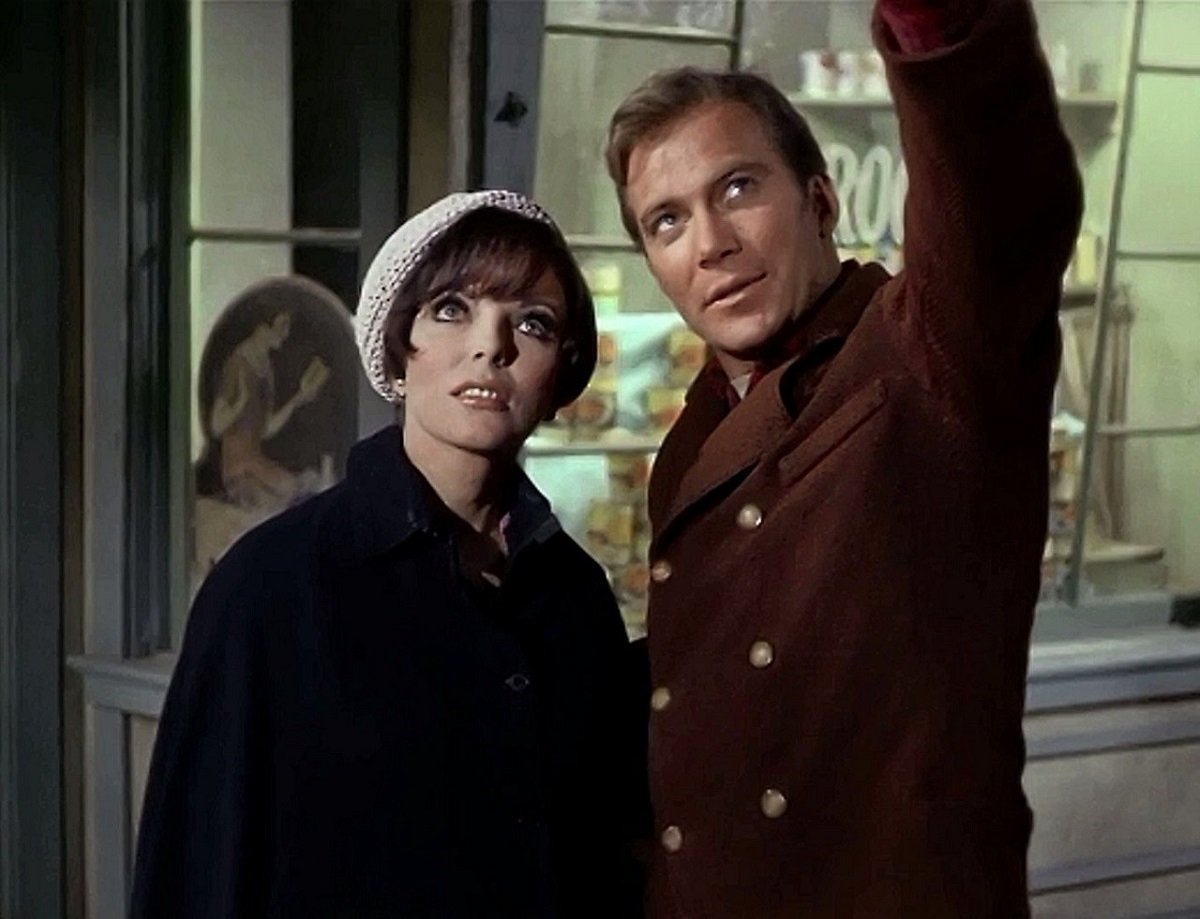 Image of Joan Collins as Edith Keeler and William Shatner as James Kirk in the STAR TREK Original Series episode, "City on the Edge of Forever." They stand on the street at night outside a store. Edith is a white woman with short dark hair and bangs under a white, knit tam hat. She's wearing a black autumn coat. Kirk is a white man slightly taller than her with short, brown hair and wearing a reddish-brown coat with gold buttons. They're both looking up at the sky as he points up toward something.