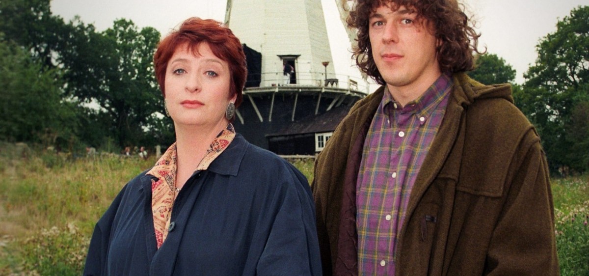 A red haired white woman stands next to a white man with long dark curls in front of a white painted windmill.