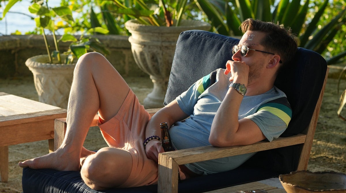 Kieran Culkin as Roman Roy in a scene from HBO's 'Succession.' He is lounging outside in the sunshine on a patio area surrounded by green plants and sitting in a patio lounger. He's a white man with dark hair wearing sunglasses, a light blue t-shirt and pastel orange shorts. He's holding a beer. 