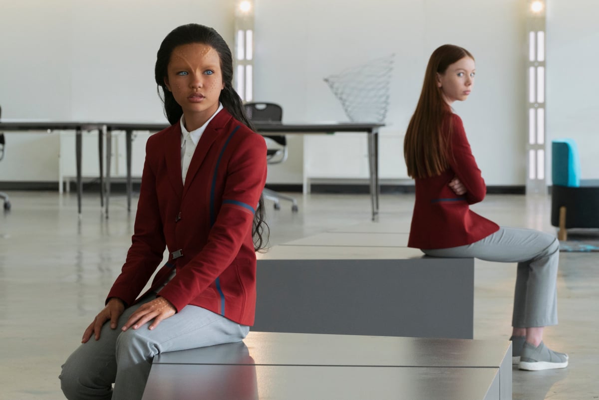 Image of Ilamaria Ebrahim as Kima and Sadie Munroe as Lil in the SHORT TREKS episode, "Children of Mars." They are both wearing the same school uniform of a red blazer, white buttondown, and grey pants with grey shoes. Kima is an alien with bronze skin, bright blue eyes, and forehead ridges. She has long, black hair. Lil is a human white girl, and has long, red hair. They are sitting on separate benches in their school's lobby - Kima in the foreground, Lil on the bench behind her. They're sitting not facing each other. 