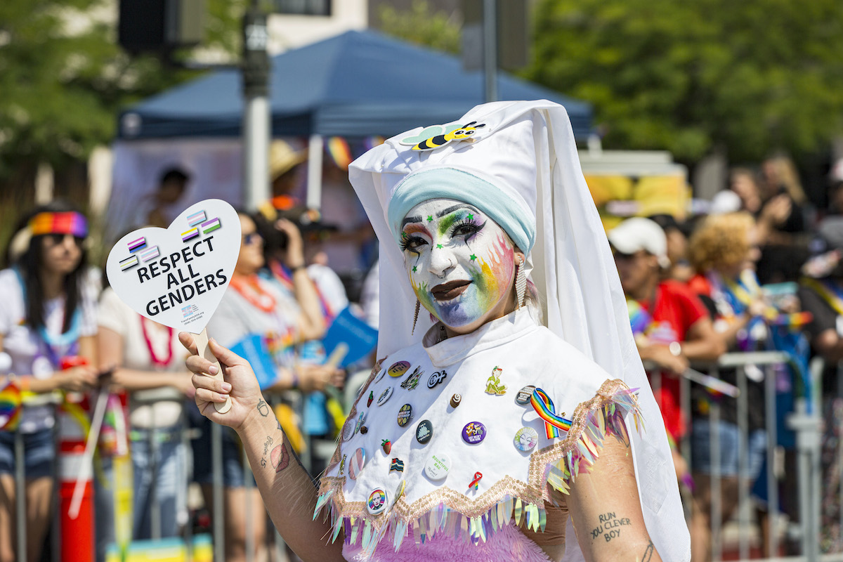 A person dressed in a white nun's habit holds a sign shaped like a heart reading "respect all genders" during a Pride parade.