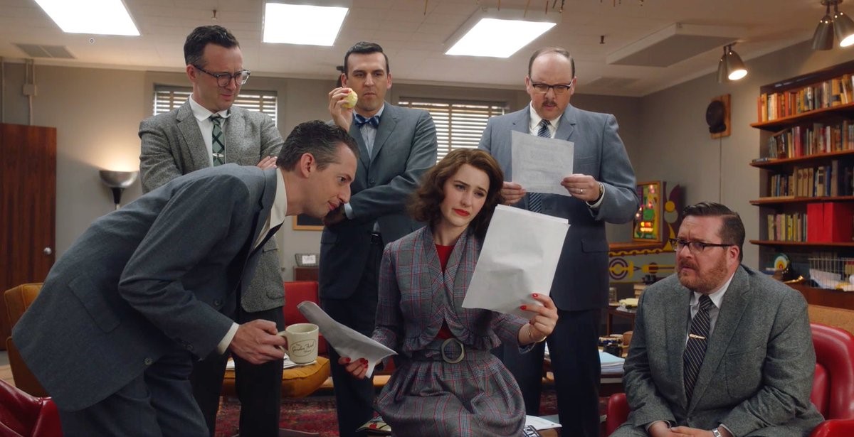Image of Rachel Brosnahan as Midge Maisel surrounded by five white men in suits in a scene from 'The Marvelous Mrs. Maisel' on Amazon. They are the writers room for the 'Gordon Ford Show." Midge, a white, Jewish woman with wavy, chin-length hair and wearing a grey skirt suit with red stripes and a red blouse under the jacket is looking at script pages skeptically as the men look at them over her shoulder. 