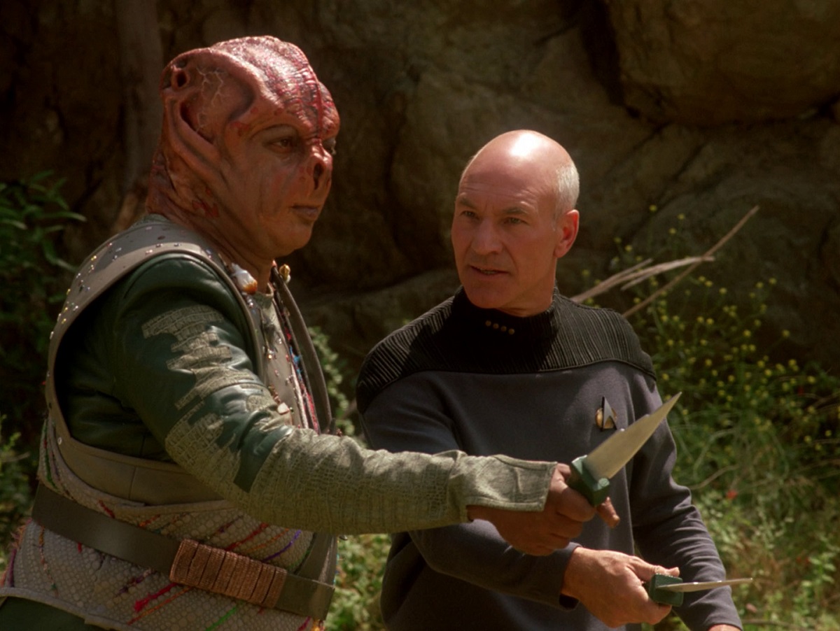 Image of Paul Winfield as Dathon and Patrick Stewart as Jean-Luc Picard in a scene from the STAR TREK: TNG episode, "Darmok." Dathon is a Tamarian, a humanoid species with reddish-brownish skin, forehead ridges, ear slits, and large, flattened nose holes. He's wearing a decorated grey vest over a greenish uniform and holding a knife out at a threat. Picard is a white man who is bald save for some white hair around the back of his head. He's wearing a grey Starfleet uniform and is also holding a knife pointed at the same threat, but he's looking to Dathon for guidance. They are outdoors.