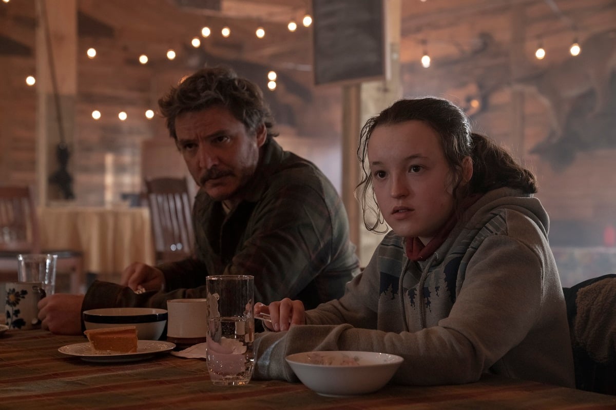 Image of Pedro Pascal as Joel and Bella Ramsay as Ellie in a scene from HBO's 'The Last of Us.' Joel is a light-skinned Latino with shaggy salt-and-pepper brown hair, a mustache and stubble and wearing a green plaid buttondown. Ellie is a white teenage girl with her brown hair pulled back into a ponytail and wearing a grey hooded sweatshirt with a red shirt underneath. They are sitting at a table eating and both looking up at something off camera. 