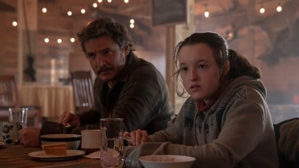 Image of Pedro Pascal as Joel and Bella Ramsay as Ellie in a scene from HBO's 'The Last of Us.' Joel is a light-skinned Latino with shaggy salt-and-pepper brown hair, a mustache and stubble and wearing a green plaid buttondown. Ellie is a white teenage girl with her brown hair pulled back into a ponytail and wearing a grey hooded sweatshirt with a red shirt underneath. They are sitting at a table eating and both looking up at something off camera.