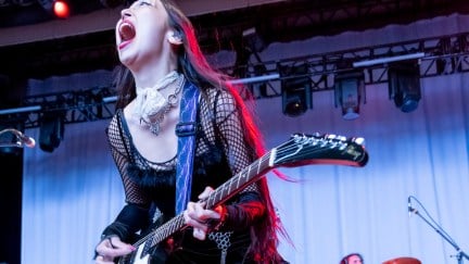 Sasami (an Asian female musician) screams into a microphone on stage while playing electric guitar.