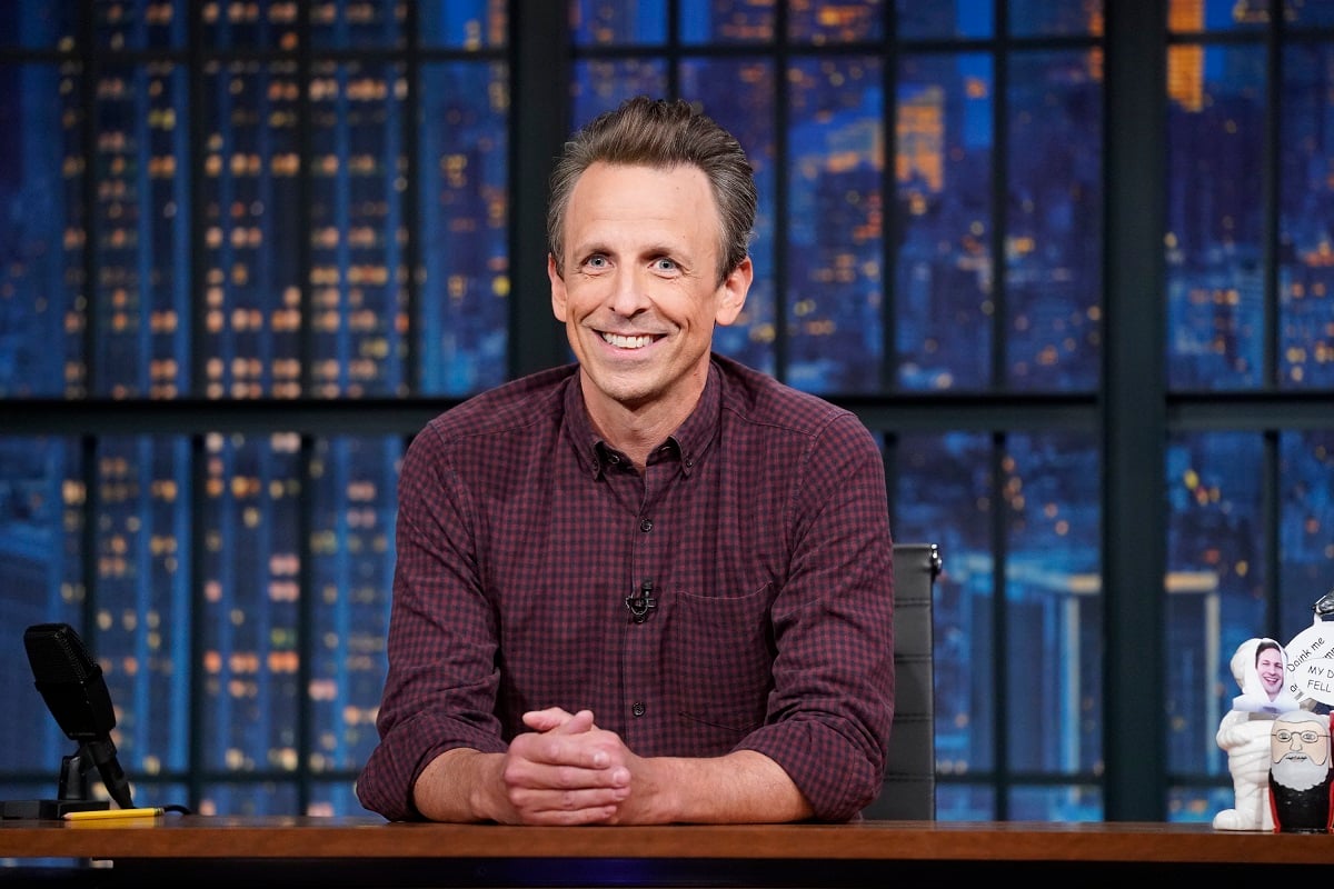 Image of Seth Meyers on the set of NBC's 'Late Night with Seth Meyers.' He is a white man with short, brown hair wearing a maroon gingham buttondown shirt. He's seated at a wooden desk with a background depicting the New York City skyline. 