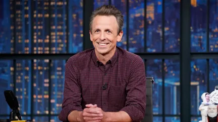 Image of Seth Meyers on the set of NBC's 'Late Night with Seth Meyers.' He is a white man with short, brown hair wearing a maroon gingham buttondown shirt. He's seated at a wooden desk with a background depicting the New York City skyline.