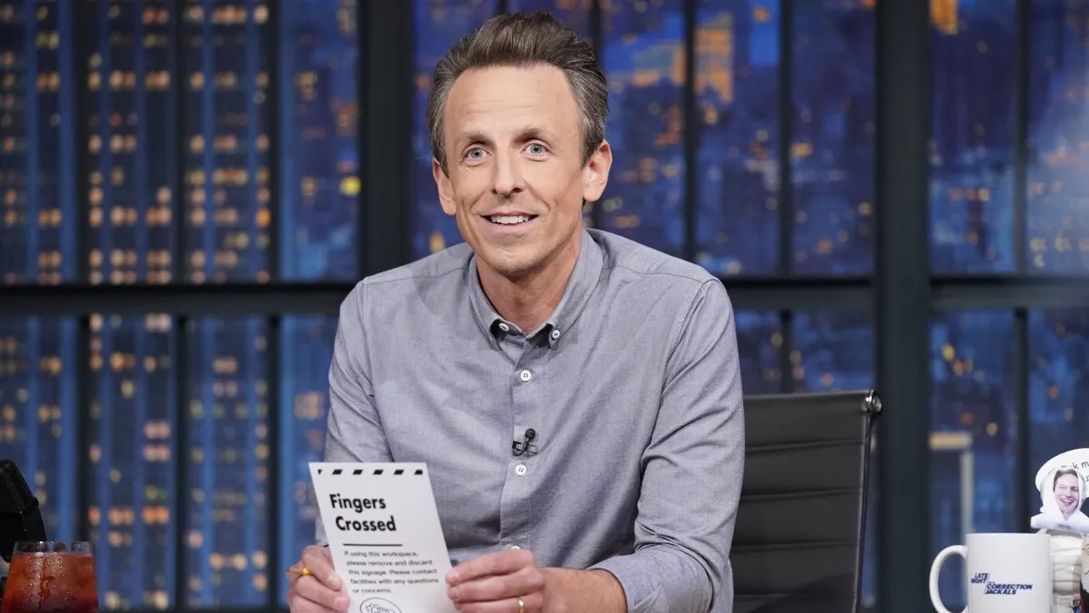 Image of Seth Meyers at his desk on the set of 'late night with Seth Meyers' on NBC. He is a white man with light brown hair and blue eyes. He's wearing a grey buttondown shirt and holding a piece of paper that has a bolded heading that reads "Fingers Crossed."