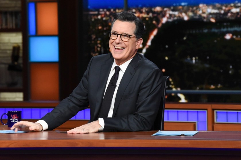 Image of Stephen Colbert on the set of CBSs 'The late show.' He is a white man with short, dark hair wearing a black suit with a white buttondown shirt and a black tie. He's seated at a wooden desk with a background depicting the New York City at night. 