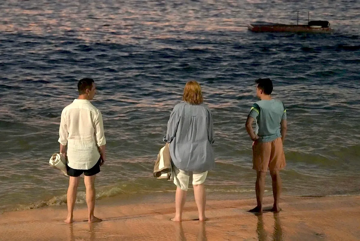 Image of Jeremy Strong as Kendall Roy, Sarah Snook as Shiv Roy, and Kieran Culkin as Roman Roy in a scene from HBO's 'Succession.' They are standing on a beach at the water's edge with their backs turned to the camera. Each are barefoot in shorts and a shirt. Kendall and Shiv are holding beach towels. It is evening.