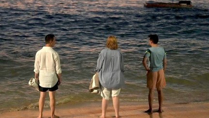 Image of Jeremy Strong as Kendall Roy, Sarah Snook as Shiv Roy, and Kieran Culkin as Roman Roy in a scene from HBO's 'Succession.' They are standing on a beach at the water's edge with their backs turned to the camera. Each are barefoot in shorts and a shirt. Kendall and Shiv are holding beach towels. It is evening.