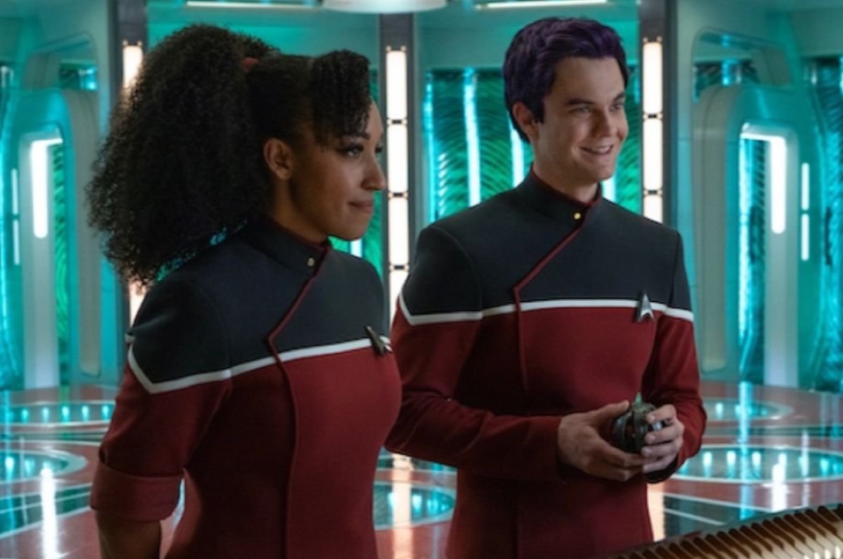 Tawny Newsome as Beckett Mariner and Jack Quaid as Bradward Boimler in a live-action scene from 'Strange New Worlds.' Beckett is a Black woman with long, curly hair tied back in a ponytail with curly bangs and wearing a red Starfleet uniform in the style of 'Lower Decks.' Brad is a white man with dyed purple hair also wearing a similar uniform. They are standing in a transporter room.