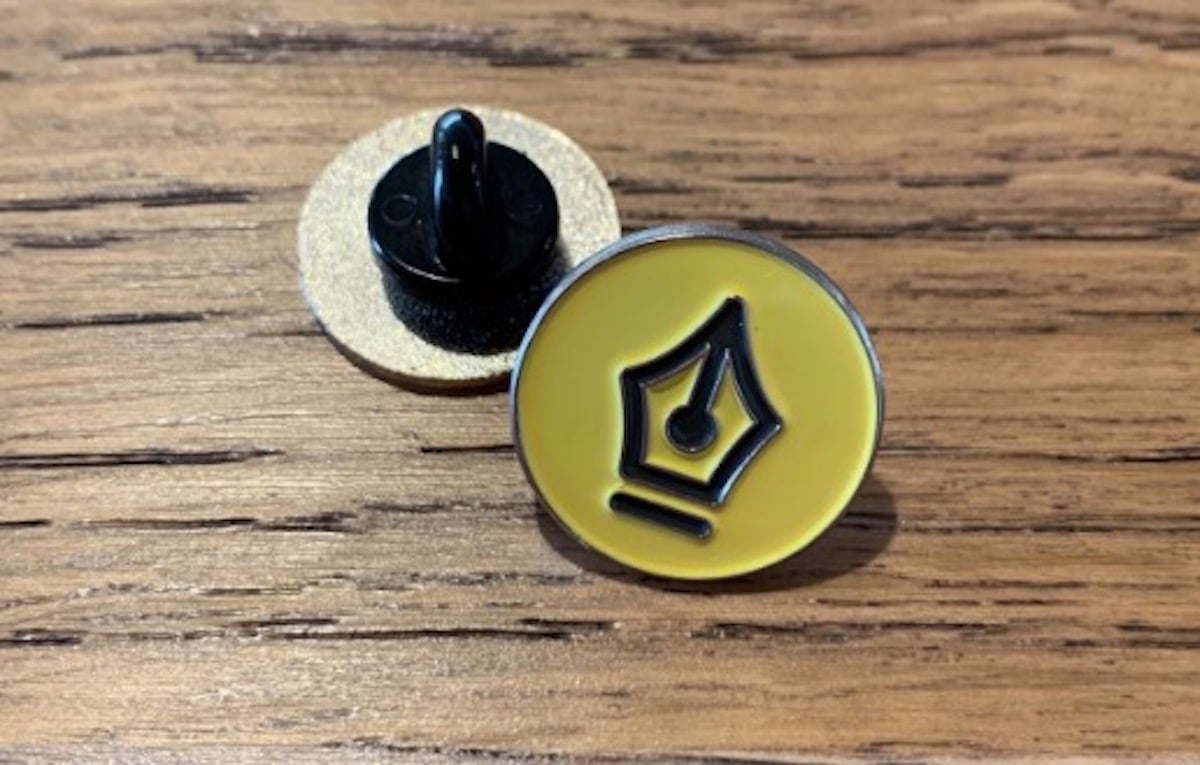 A small yellow pin featuring the logo for The Nib, the tip of an ink fountain pen.