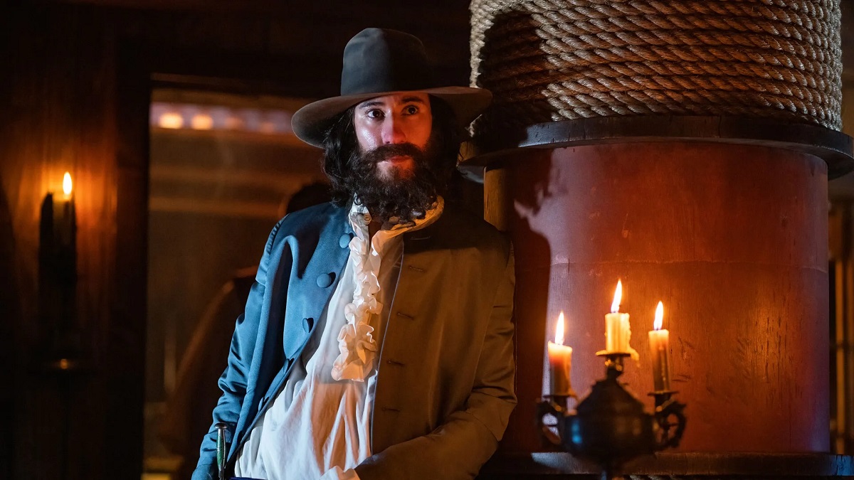 Image of Vico Ortiz as Jim on HBO Max's 'Our Flag Means Death.' Jim is a Latine, masc, non-binary person leaning against the mast in a ship cabin. They're wearing a wide-brimmed hat, have shoulder-length black hair and a curly beard, and are wearing a bluish, early 18th Century period jacket with a white shirt with a frilly collar underneath. There's a lit candelabra beside them.