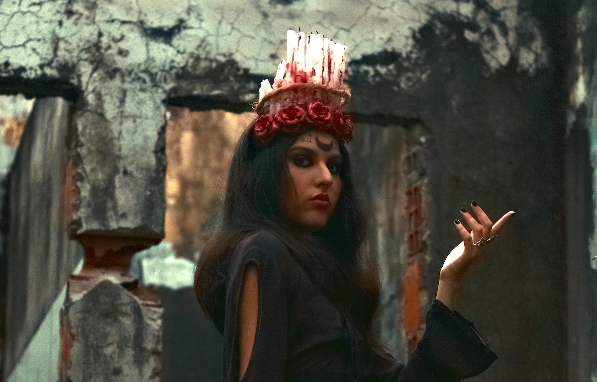 A woman with a black moon on her forehead and a headdress made of candles poses against a crumbling concrete structure.