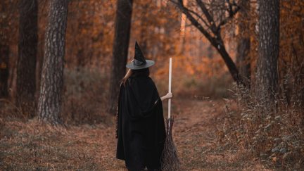 A figure in a black cloak, black witch's hat, and broom walks through the forest.