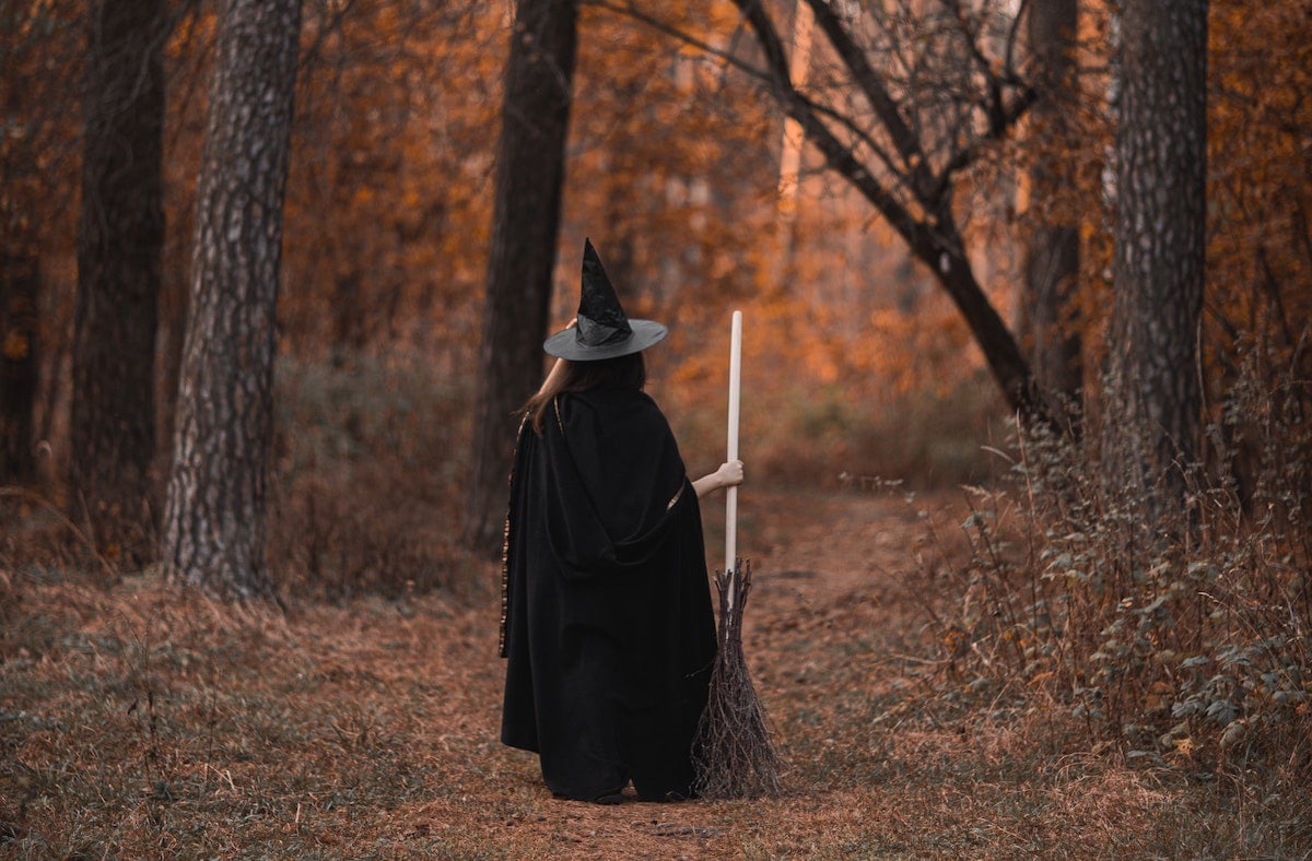 A figure in a black cloak, black witch's hat, and broom walks through the forest.