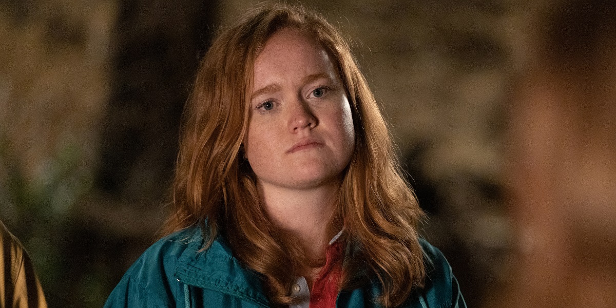 Image of Liv Hewson as Val in a scene from 'Yellowjackets' on Showtime. Val is a white teenage girl with long, red hair wearing a blue-green jacket over a red collared shirt. She's standing outside. 