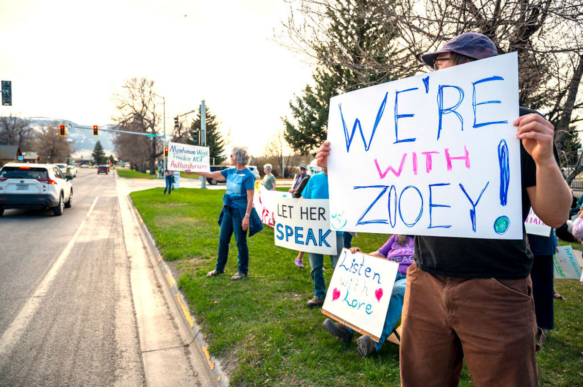 People stand by the side of a road holding signs that say "We're with Zooey!" and "Let her speak".