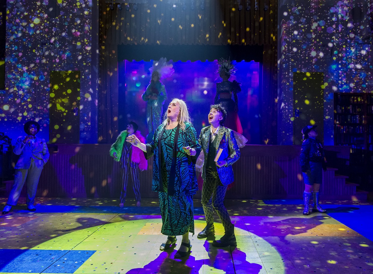 Image from 'A Transparent Musical' at the Mark Taper Forum in L.A. Maura and Ari stand in the middle of the stage looking up dazzled by falling confetti and twinkling lights. Both are white and Jewish. Maura has long white hair and is wearing a green dress with a leaf pattern. Ari has short curly dark hair and is wearing a patterned blazer, a dark vest over a light buttondown, and dark pants with a criss-cross pattern.