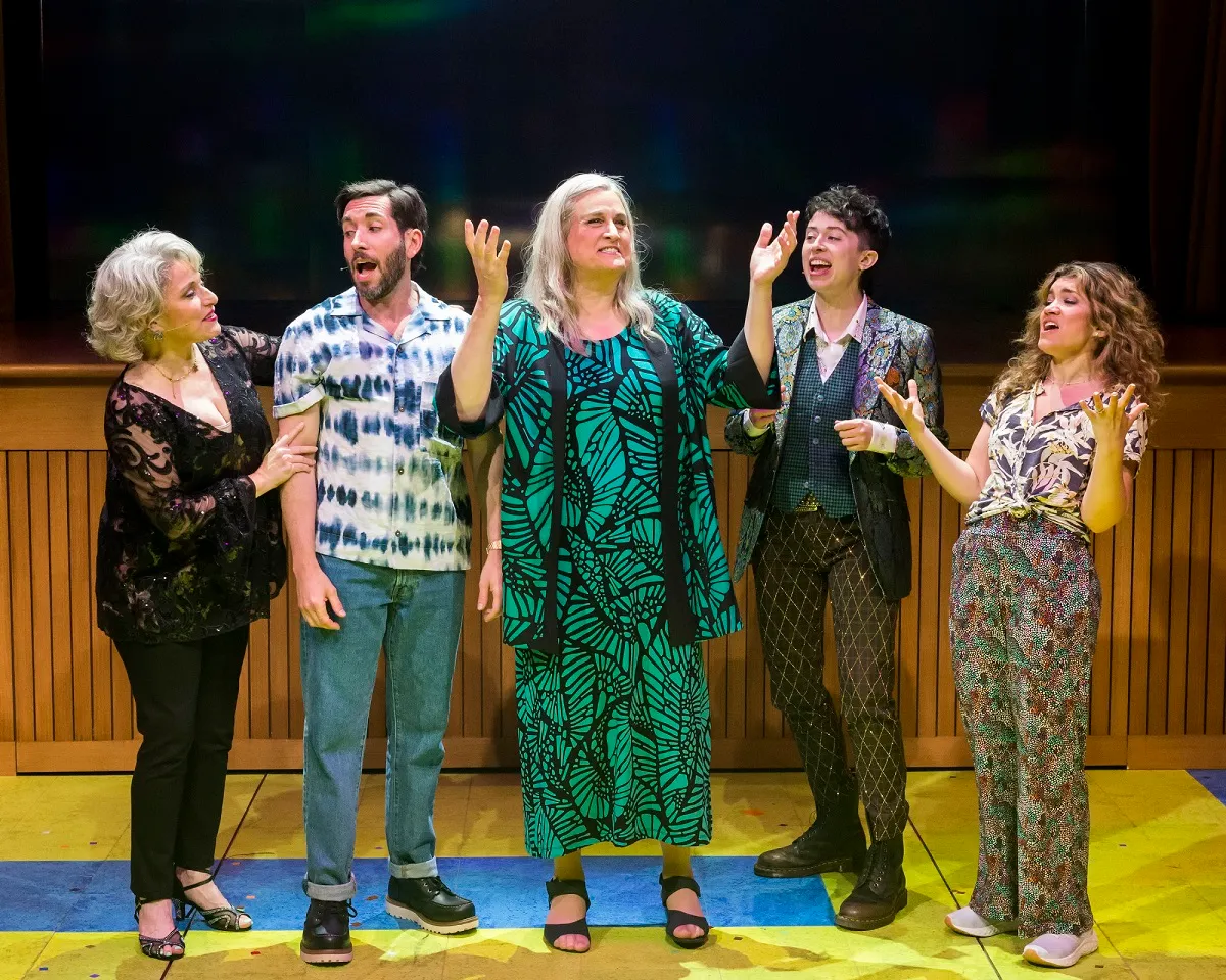 Image from 'A Transparent Musical' at the Mark Taper Forum in L.A. The Pfefferman family stand in a row, all of them white and Jewish. Shelley (short with white hair and wearing all black), Josh, (dark hair and beard wearing a patterned buttondown and jeans), Maura (long white hair and wearing a green dress with leaf patterns), Ari (short, curly dark hair wearing a patterned blazer, a dark vest over a light buttondown, and dark pants with a criss-cross pattern), and Sarah (long curly brown hair and wearing a plant print buttondown and patterned flowy pants).