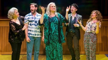 Image from 'A Transparent Musical' at the Mark Taper Forum in L.A. The Pfefferman family stand in a row, all of them white and Jewish. Shelley (short with white hair and wearing all black), Josh, (dark hair and beard wearing a patterned buttondown and jeans), Maura (long white hair and wearing a green dress with leaf patterns), Ari (short, curly dark hair wearing a patterned blazer, a dark vest over a light buttondown, and dark pants with a criss-cross pattern), and Sarah (long curly brown hair and wearing a plant print buttondown and patterned flowy pants).