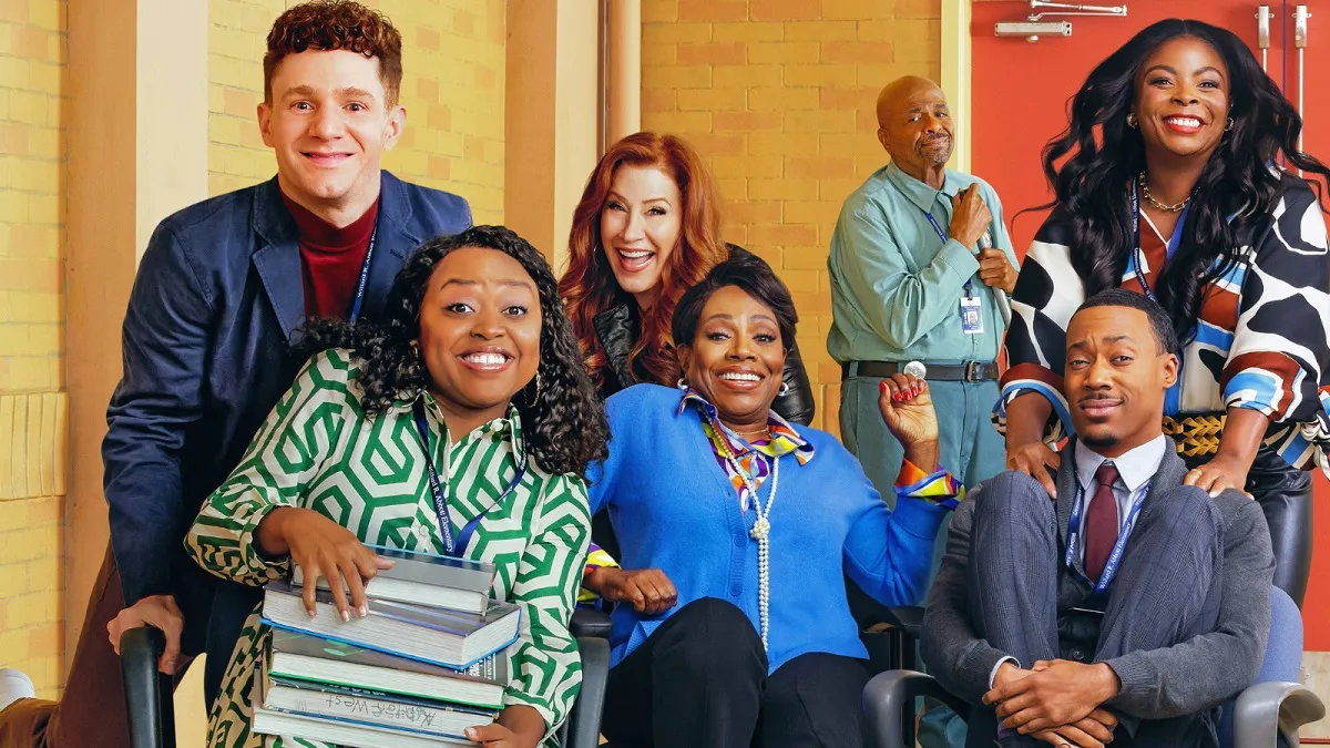 The cast of ABC's 'Abbott Elementary.' Jacob (white man - played by Chris Perfetti, Janine (Black woman - played by Quinta Brunson), Melissa (white woman - played by Lisa Ann Walter), Barbara (Black woman - played by Sheryl Lee Ralph), Gregory (Black man - played by Tyler James Williams), and Ava (Black woman - played by Janelle James) sit/stand around each other in a huddle in a school hallway as janitor Mr. Johnson (Black man - played by William Stanford Davis) looks on in the background.
