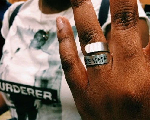 A Black woman holds out her hand, on it is a silver wrap around ring with the word "femme" stamped on it.
