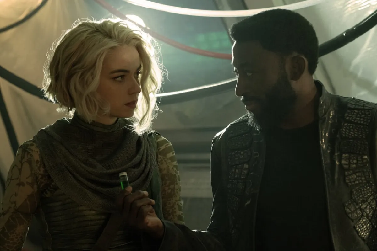 Chapel (Jess Bush) and M'Benga (Babs Olusanmokun) in a scene from 'Star Trek: Strange New Worlds.' They look at each other as M'Benga hands Chapel a vial with a green liquid in it. Chapel is a blonde, white woman with a chin-length, wavy bob wearing a light green cowl and a patterned, tight, green, long-sleeved shirt. M'Benga is a Black man with short, dark hair and a beard wearing a black jacket with reptile-textured accents and a black shirt.