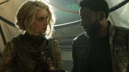 Chapel (Jess Bush) and M'Benga (Babs Olusanmokun) in a scene from 'Star Trek: Strange New Worlds.' They look at each other as M'Benga hands Chapel a vial with a green liquid in it. Chapel is a blonde, white woman with a chin-length, wavy bob wearing a light green cowl and a patterned, tight, green, long-sleeved shirt. M'Benga is a Black man with short, dark hair and a beard wearing a black jacket with reptile-textured accents and a black shirt.