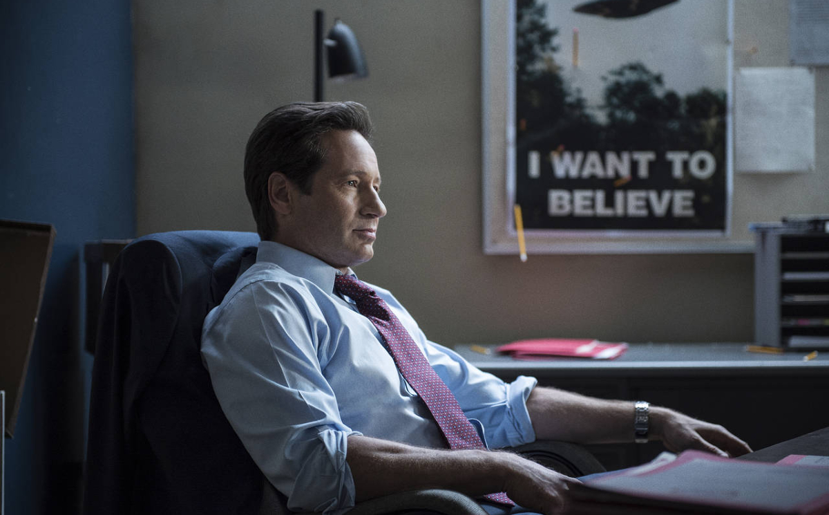 Fox Mulder (David Duchovny) sits at his desk, a poster featuring a flying saucer and the caption "I want to believe" hangs in the background