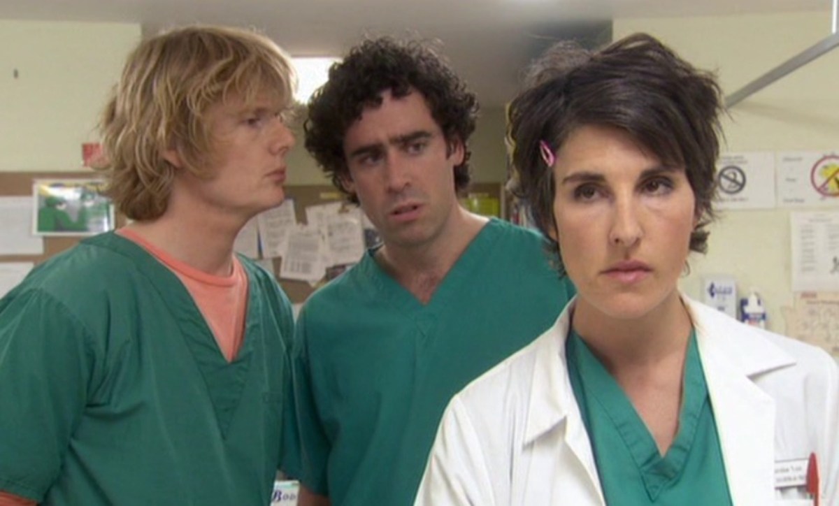 Still from Green Wing; Julian Rhind Tutt, a white man with blonde hair, stands next to Stephen Mangan, a white man with dark hair. Both wearing green scrubs. In front of them stands Tamsin Greig, a dark haired white woman, in a white doctors coat.