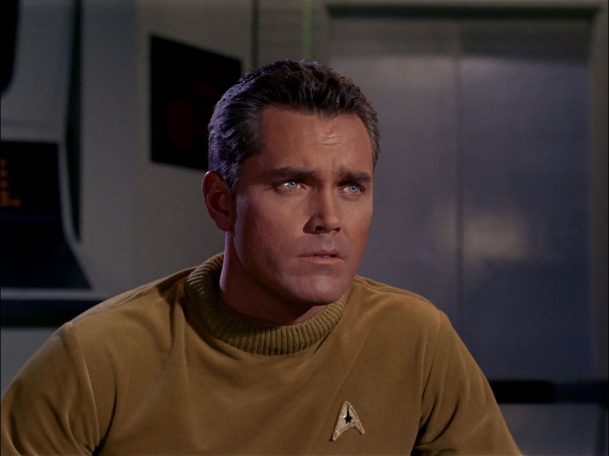 Jeffrey Hunter as Captain Christopher Pike in the unaired 'Star Trek' pilot, "The Cage." He is a white man with black hair wearing a gold Starfleet shirt. He looks concerned.