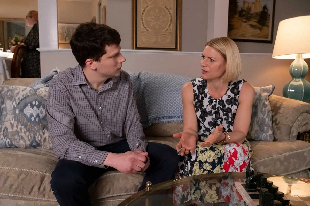 Jesse Eisenberg as Toby and Claire Danes as Rachel in Hulu's 'Fleishman is in Trouble.' They are both white and Jewish and seated on a couch at a gathering. Toby has dark hair and is wearing a grey pin-striped buttondown and dark pants. Rachel has a blonde, chin-length bob and is wearing a sleeveless black and white dress. They are mid-conversation and looking annoyed. 