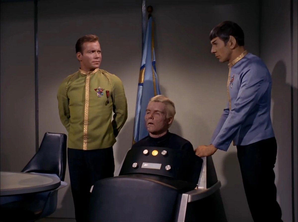 Kirk (William Shatner), Pike (Sean Kenney), and Spock (Leonard Nimoy) in a scene from 'Star Trek.' Kirk is a white man with brown hair standing at ease while wearing a gold Starfleet dress uniform jacket and black pants. Pike is in a brainwave-operated wheelchair that he's encased in up to his chest. He's a white man with white hair and a burned face. Standing with his hand on the chair, Spock is a white Vulcan with pointy ears and short, dark hair wearing a blue Starfleet dress uniform jacket and black pants. 