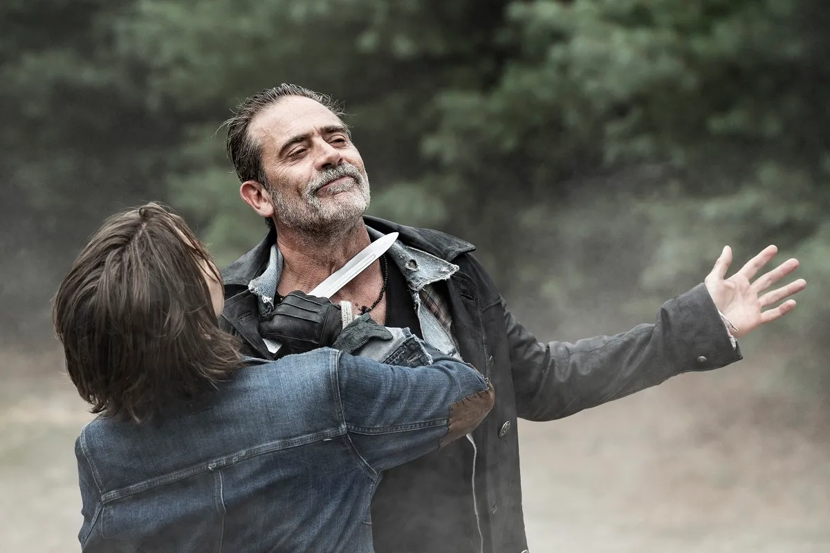 Lauren Cohan as Maggie Rhee and Jeffrey Dean Morgan as Negan in a scene from AMC's 'The Walking Dead: Dead City.' Maggie is a white woman with chin-length brown hair holding a knife to Negan's throat in her black fingerless gloves, wearing a denim jacket. Negan is a white man with dark hair and a salt-and-pepper 5 o'clock shadow wearing several layers of shirts under a black jacket. He's holding his arms out with smirk on his face accepting that a knife to his throat is how it's gonna be now. They're standing outside with grass and trees in the background.
