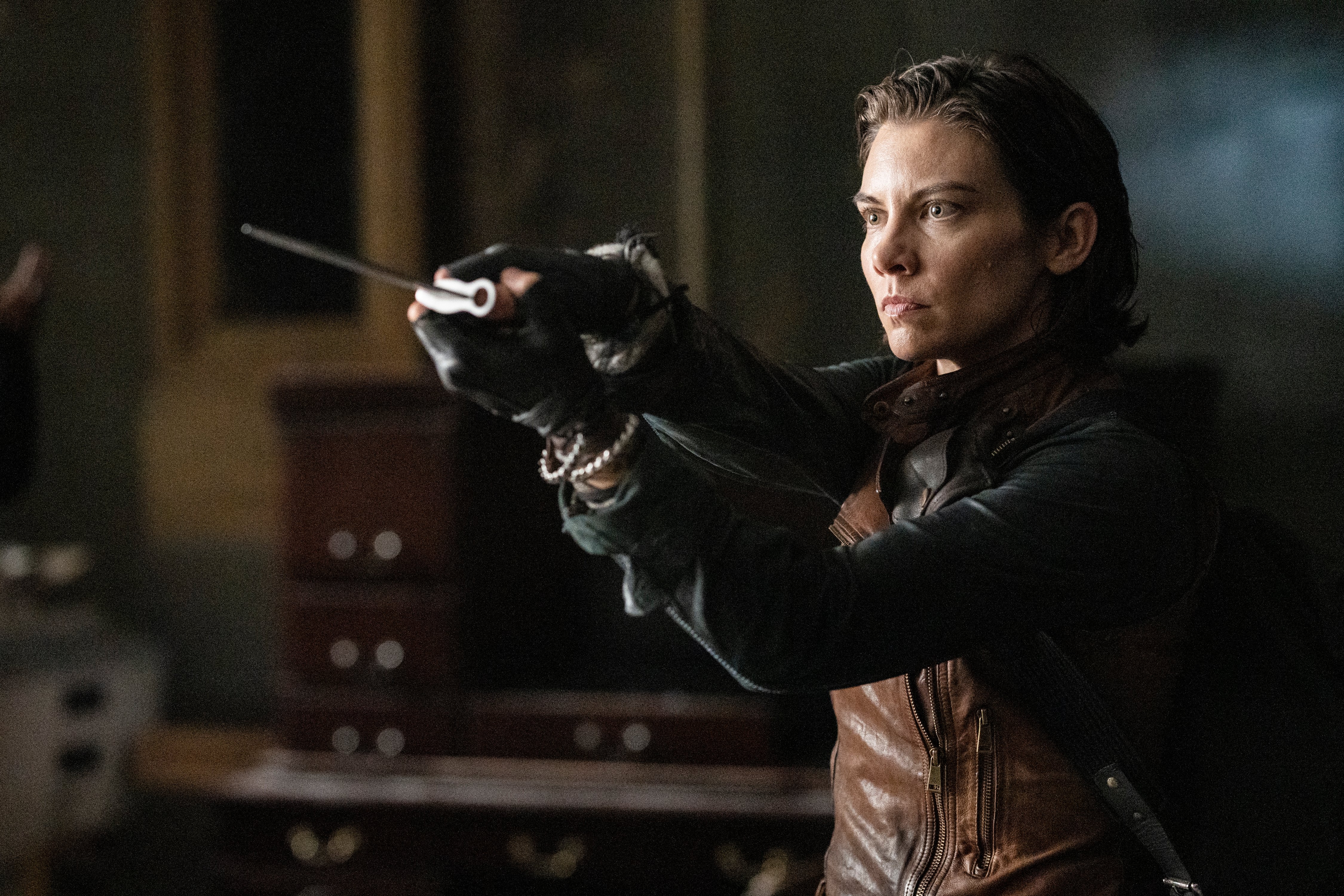 Lauren Cohan as Maggie Rhee in a scene from AMC's 'The Walking Dead: Dead City.' Maggie is a white woman with chin-length brown hair holding a knife out with both hands  in black fingerless gloves, wearing a brown, leather vest over a long-sleeved black shirt. She is indoors.