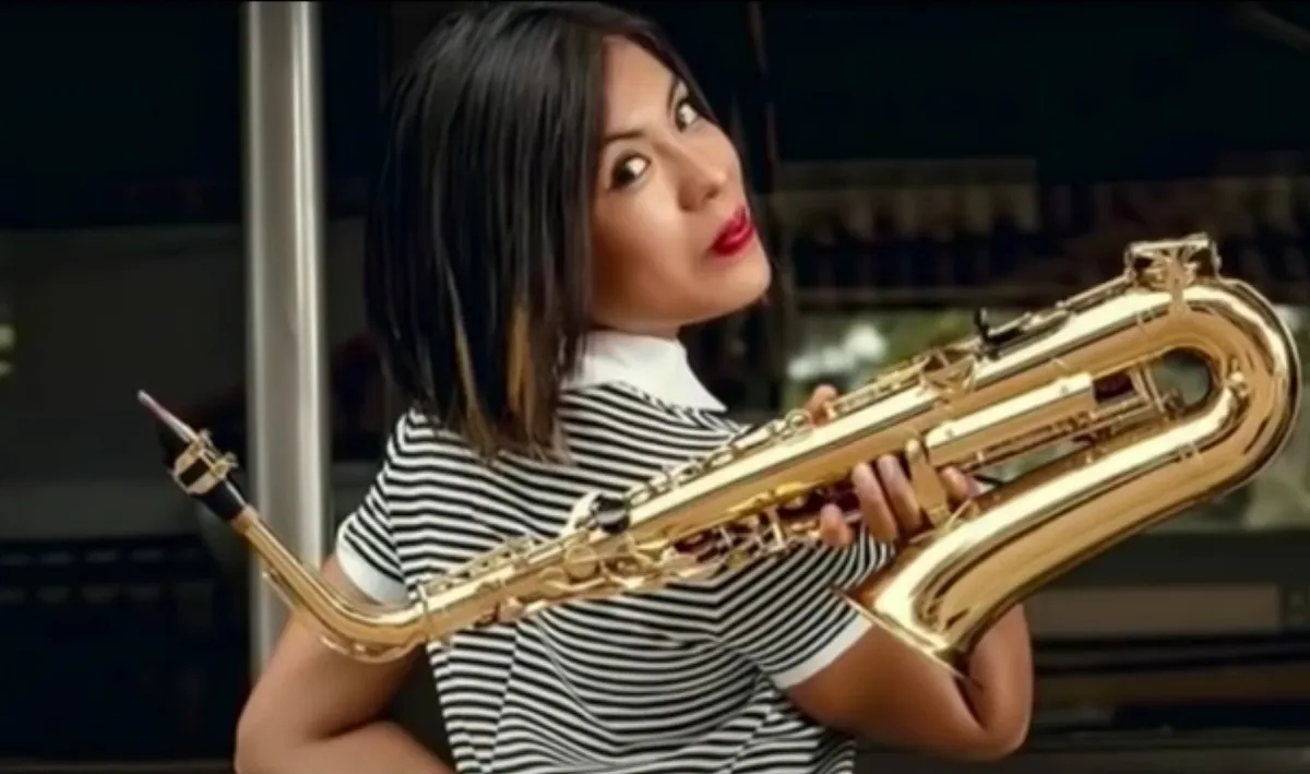 Screengrab of María Elena Ríos, a brown Mexican woman with shoulder-length dark hair with highlighted tips. She is smirking as she looks over her shoulder at the camera while holding a saxophone over her shoulder. She's wearing a black and white striped short-sleeved, collared shirt. 