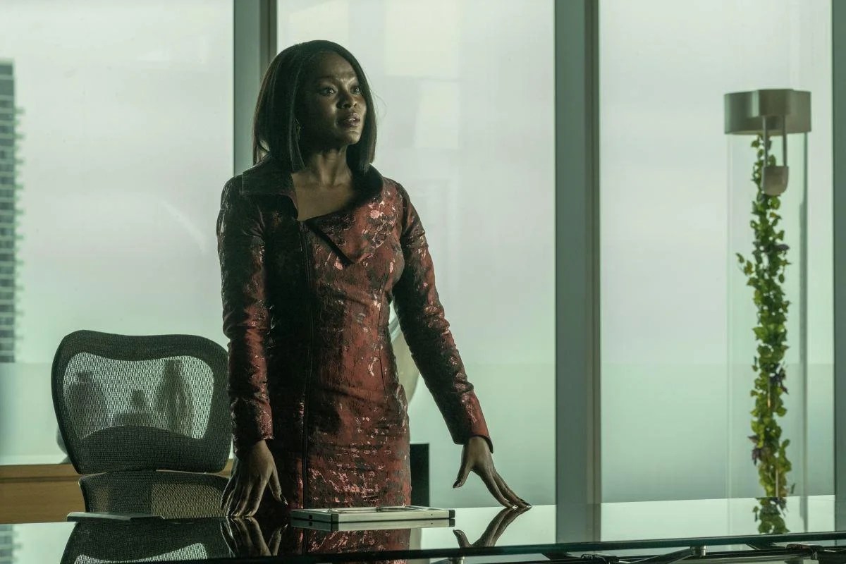 Neera (Yetide Badaki) in a scene from 'Strange New Worlds' on Paramount+. Neera is a dark-skinned Black woman with straight black hair wearing a red, iridescent dress with long sleeves and an asymetrical neckline. She is standing at a glass desk in her office with her hands on the table as she looks at someone across from her. There are floor to ceiling windows behind her.