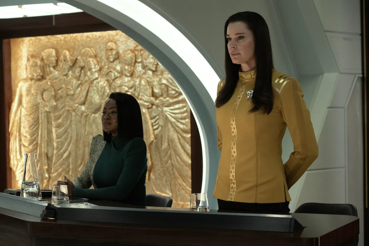 Neera (Yetide Badaki) and Una (Rebecca Romijn) in a scene from 'Strange New Worlds' on Paramount+. Neera is a dark-skinned Black woman with straight black hair wearing a blue-green dress with one white speckled sleeve and shoulder that covers her arms and neck. She is seated with her hands folded at a table in a courtroom. Una is a white woman with long, black hair wearing a gold Starfleet dress uniform. She is standing at ease at the table.
