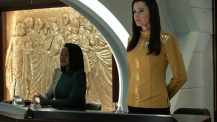 Neera (Yetide Badaki) and Una (Rebecca Romijn) in a scene from 'Strange New Worlds' on Paramount+. Neera is a dark-skinned Black woman with straight black hair wearing a blue-green dress with one white speckled sleeve and shoulder that covers her arms and neck. She is seated with her hands folded at a table in a courtroom. Una is a white woman with long, black hair wearing a gold Starfleet dress uniform. She is standing at ease at the table.