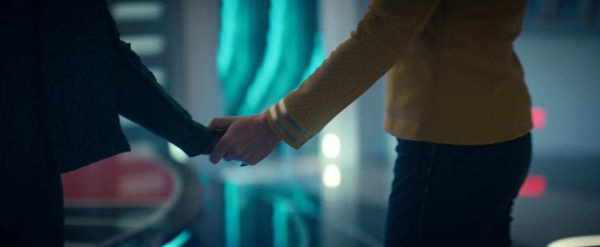 Una (Rebecca Romijn) and Neera (Yetide Badaki) in a scene from 'Strange New Worlds' on Paramount+. We see a close-up of their hands as they hold each other's hand. One a Black woman's who's wearing a dark, long-sleeved dress. The other a white woman's hand wearing a gold, long-sleeved shirt and black pants as the Black woman walks away from the white woman in the transporter room.