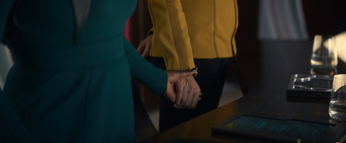 Una (Rebecca Romijn) and Neera (Yetide Badaki) in a scene from 'Strange New Worlds' on Paramount+. We see a close-up of their hands as they hold each other's hand. One a Black woman's who's wearing a blue-green dress. The other a white woman's hand wearing a gold, long-sleeved shirt and black pants as they stand together behind a desk. 